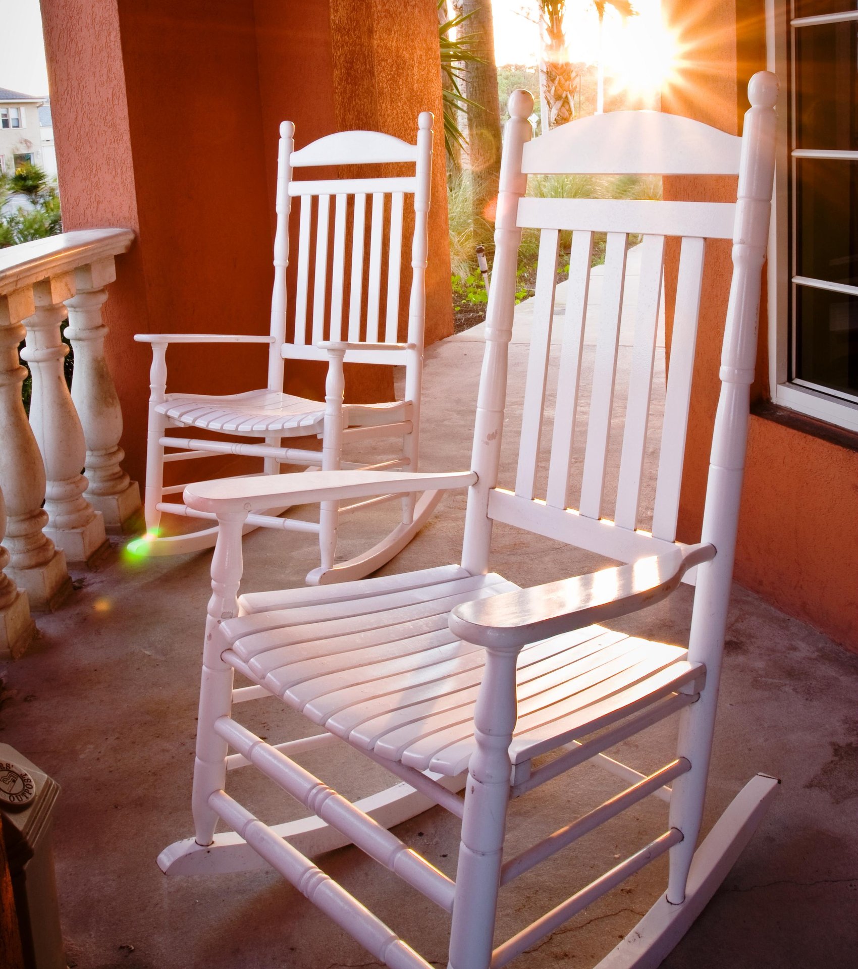 Two white wooden rocking chairs sit on the porch of Amelia Hotel at the Beach, with warm sunlight streaming across the orange walls and railing, creating a peaceful coastal atmosphere.