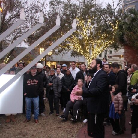 A man speaks to a large crowd gathered outdoors at dusk beside a giant menorah. Trees decorated with string lights are visible behind the crowd, and people of all ages watch the event.