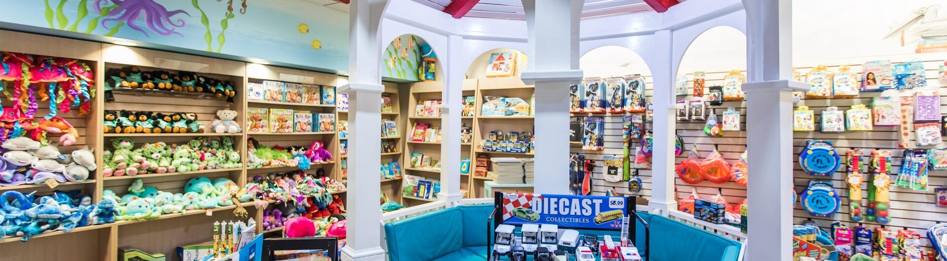 A colorful toy store with shelves full of stuffed animals, books, and various toys. A white gazebo with red trim and blue seats is in the center, and a display of die-cast toy cars is in the foreground.