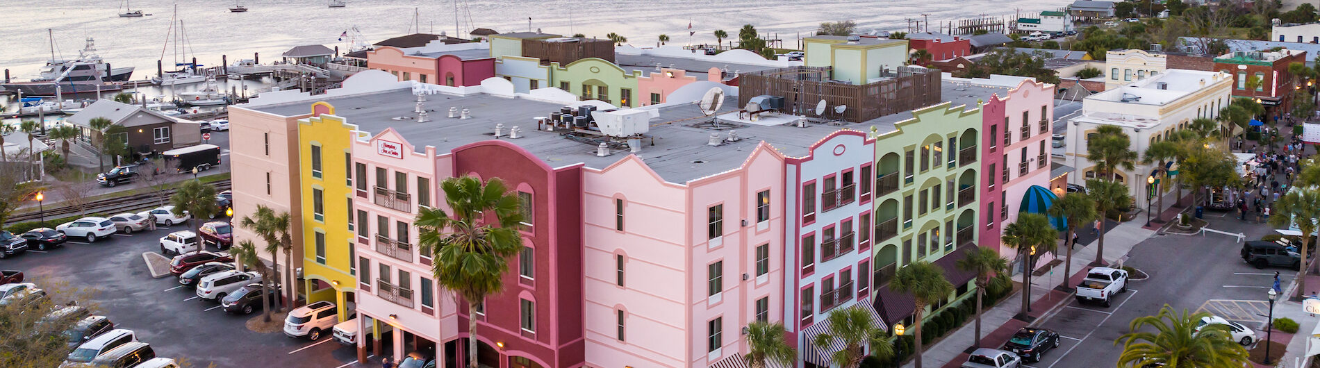 Aerial view of pastel-colored buildings and palm trees along a street near a marina at sunset, with cars parked and boats docked by the waterfront.