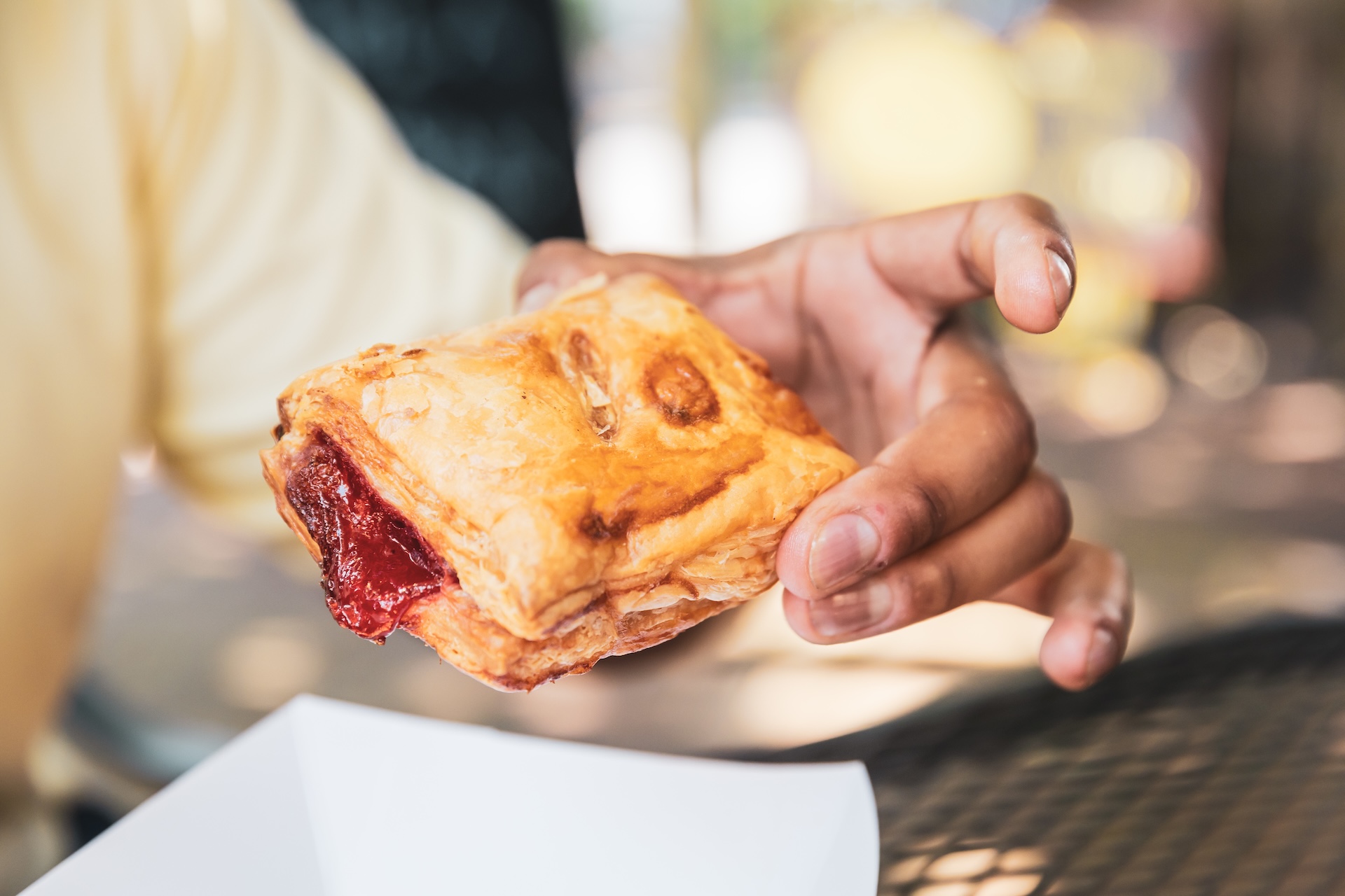 A person holding a rectangular pastry with golden, flaky crust and red filling, possibly jam or fruit, over a white tray. The background is blurred, showing an outdoor setting.