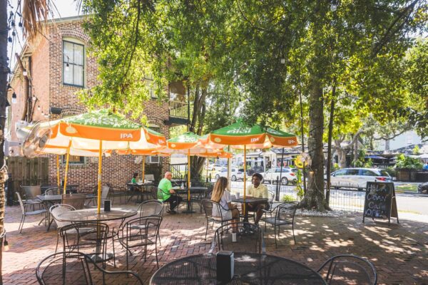 A sunny outdoor café patio with metal tables and chairs, green and orange umbrellas, and people sitting and chatting under leafy trees. A chalkboard sign stands by the entrance, and cars are parked along the street beyond the patio.