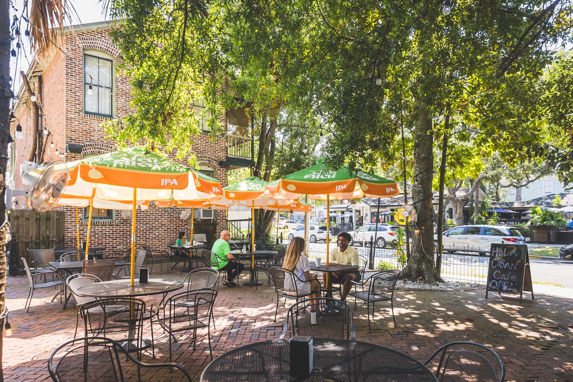 A sunny outdoor café patio with metal tables and chairs, green and orange umbrellas, and people sitting and chatting under leafy trees. A chalkboard sign stands by the entrance, and cars are parked along the street beyond the patio.