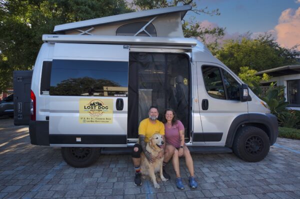 A smiling man and woman sit with a golden retriever in front of a silver camper van with a Lost Dog coffee shop logo. The van is parked on a cobblestone driveway surrounded by greenery, under a partly cloudy sky.