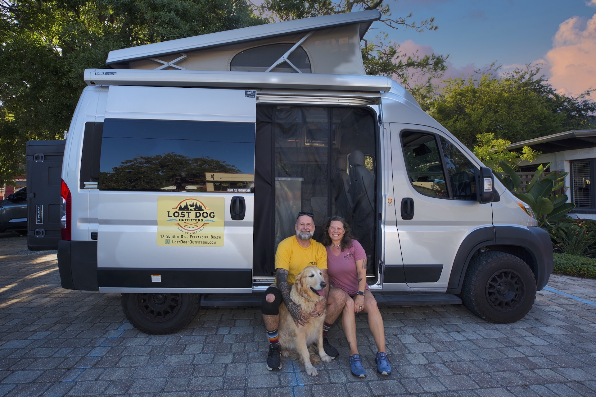 A smiling man and woman sit with a golden retriever in front of a silver camper van with a Lost Dog coffee shop logo. The van is parked on a cobblestone driveway surrounded by greenery, under a partly cloudy sky.