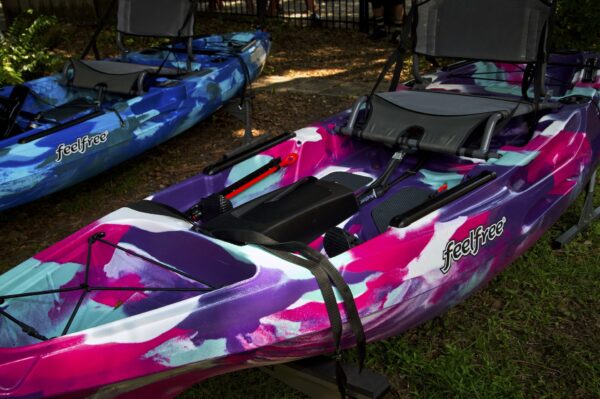 A close-up of a colorful kayak with vibrant pink, purple, and blue patterns, featuring the brand name feelfree. Another blue kayak is visible in the background, both resting on stands outdoors.