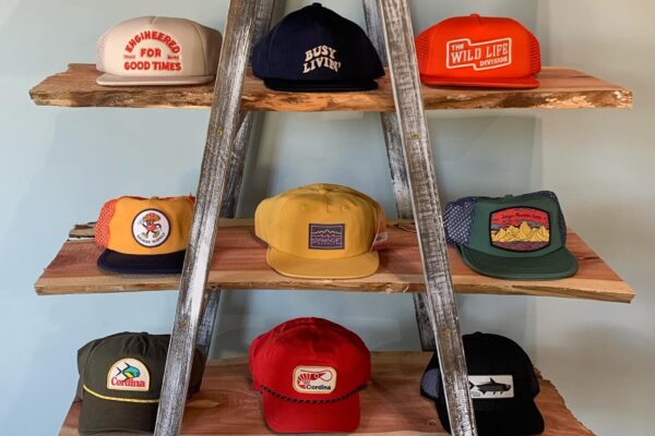 Nine colorful trucker hats with various patches and designs are displayed on three wooden shelves supported by a rustic wooden ladder against a light-colored wall.