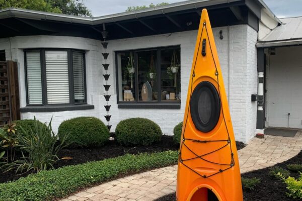 An orange kayak stands upright on a brick walkway in front of a white house with large windows, trimmed bushes, and hanging plants.