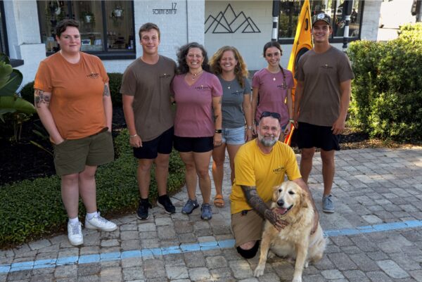 Seven people and a Golden Retriever pose outdoors on a paved area in front of a building. Six people are standing, one man is kneeling with the dog. They are all smiling and wearing casual clothes.