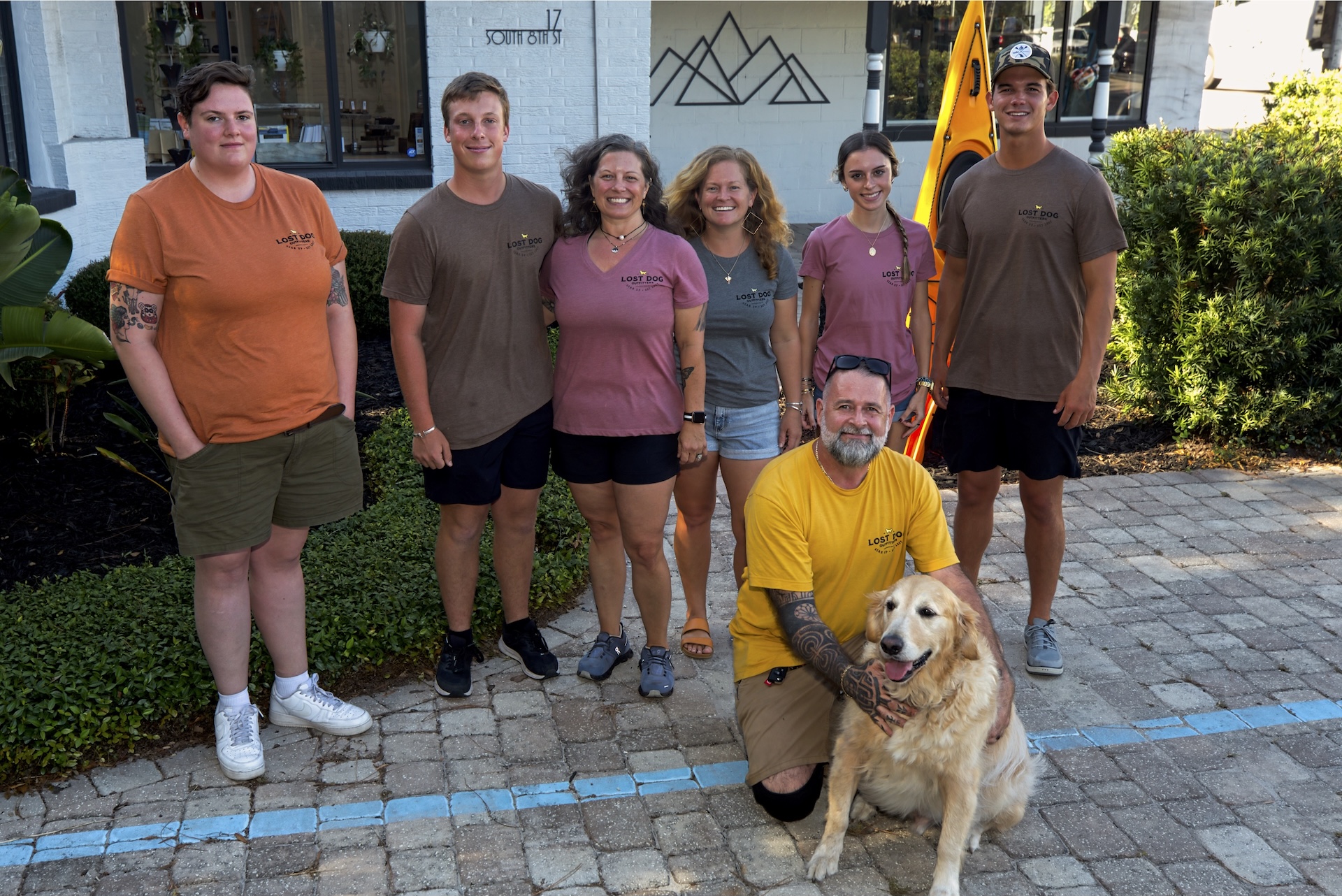 Seven people and a Golden Retriever pose outdoors on a paved area in front of a building. Six people are standing, one man is kneeling with the dog. They are all smiling and wearing casual clothes.