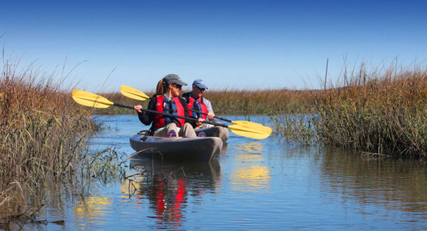 Two people wearing life jackets and hats paddle a tandem kayak with yellow oars through calm marsh waters surrounded by tall grasses under a bright blue sky.