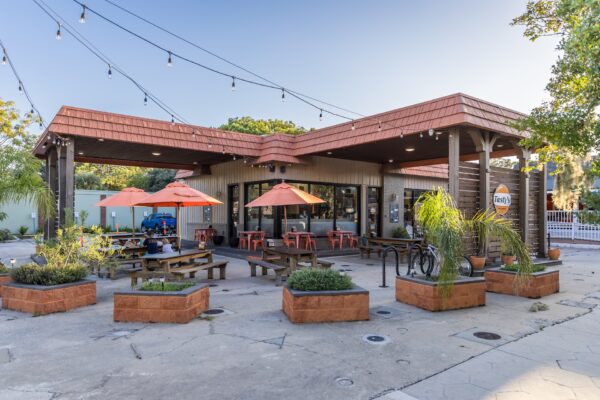 A modern cafe with a large outdoor patio, orange umbrellas, wooden picnic tables, string lights overhead, and potted plants in front of the building on a sunny day.