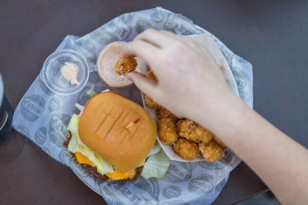 A hand dips a tater tot into a sauce next to a serving of more tater tots and a cheeseburger with lettuce and tomato, all on a tray lined with printed paper.