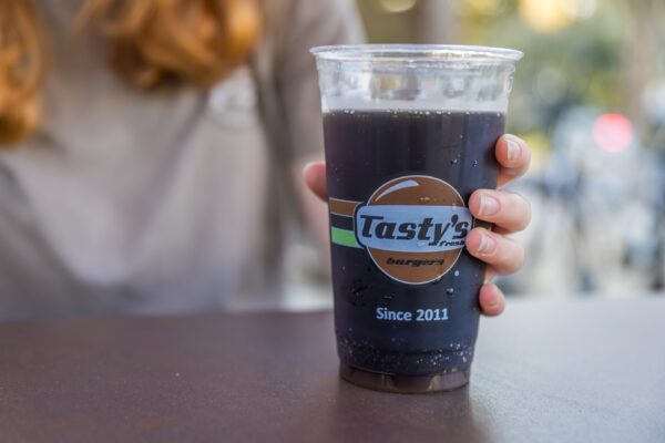 A person with long, reddish hair holds a large plastic cup of dark soda on a table. The cup features the Tastys Fresh Burgers Since 2011 logo. The background is blurred.