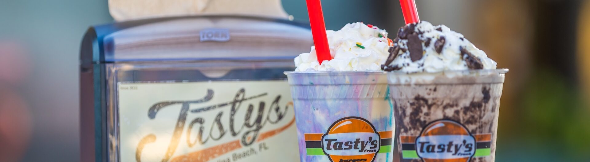 Two milkshakes topped with whipped cream and red straws sit on a table beside a napkin dispenser with the Tasty’s logo. The background is blurred.