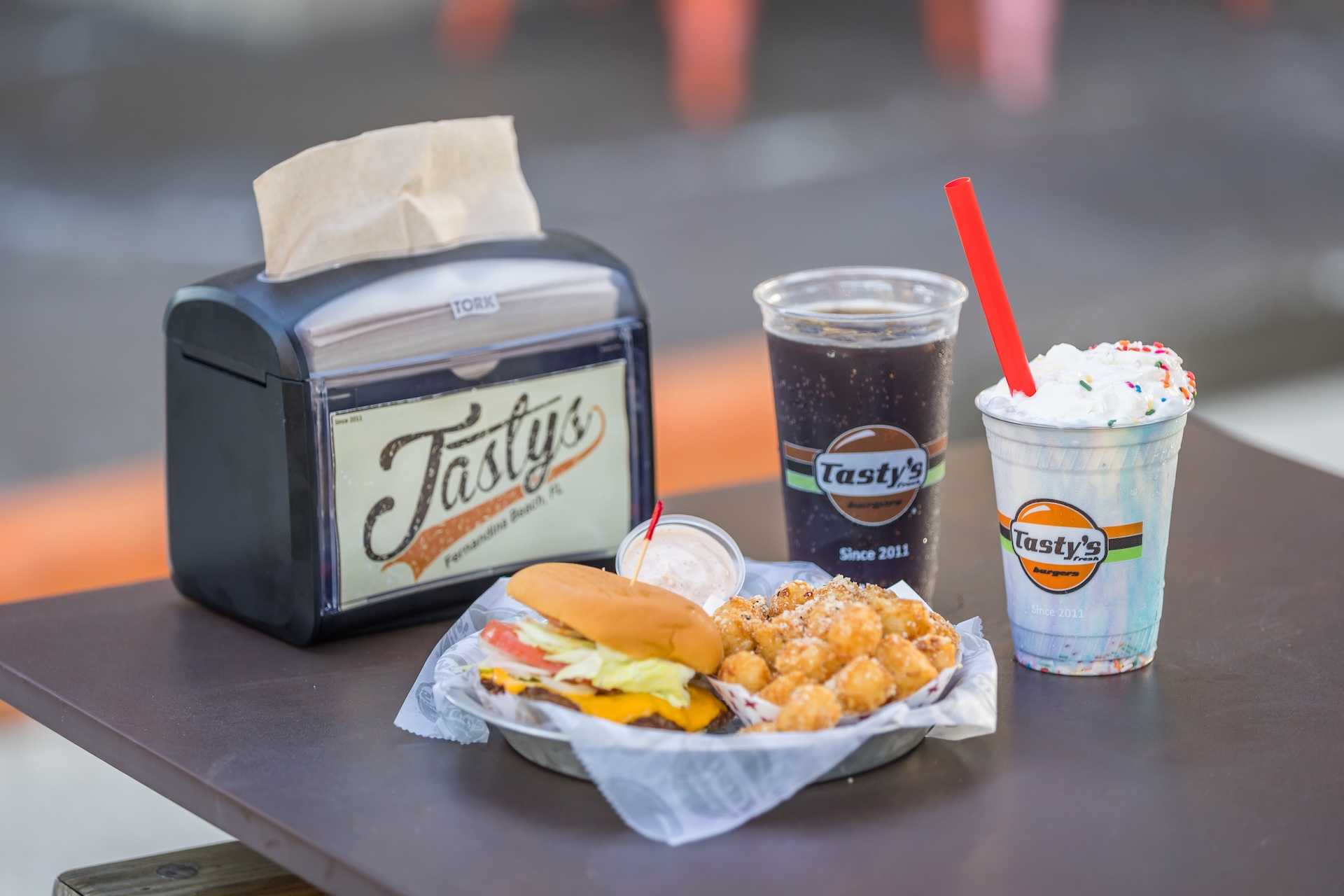A tray with a cheeseburger, tater tots, a cup of soda, and a milkshake with whipped cream and sprinkles sits on a table next to a napkin dispenser labeled Tastys.