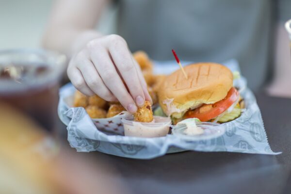 A person dips a fried chicken nugget into sauce next to a tray holding a sandwich with lettuce, tomato, and pickles, and more nuggets.