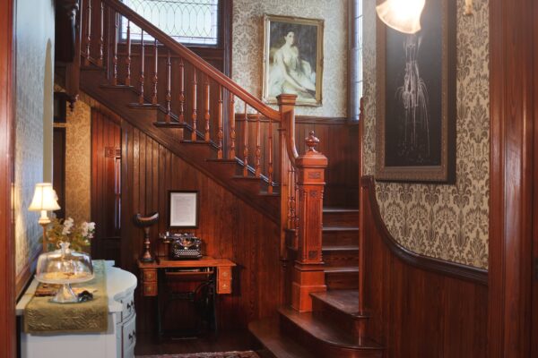 An elegant, vintage wooden staircase with ornate banisters leads to an upper floor. An old typewriter and gramophone sit on a desk beneath the stairs. Classic wallpaper and framed art decorate the walls.