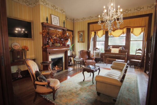 Elegant vintage living room with ornate fireplace, antique furniture, chandelier, large windows with yellow curtains, a piano, and fall-themed decorations, creating a warm and inviting atmosphere.